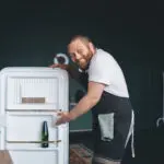 Smiling man standing next to an open refrigerator, pointing inside the empty shelves